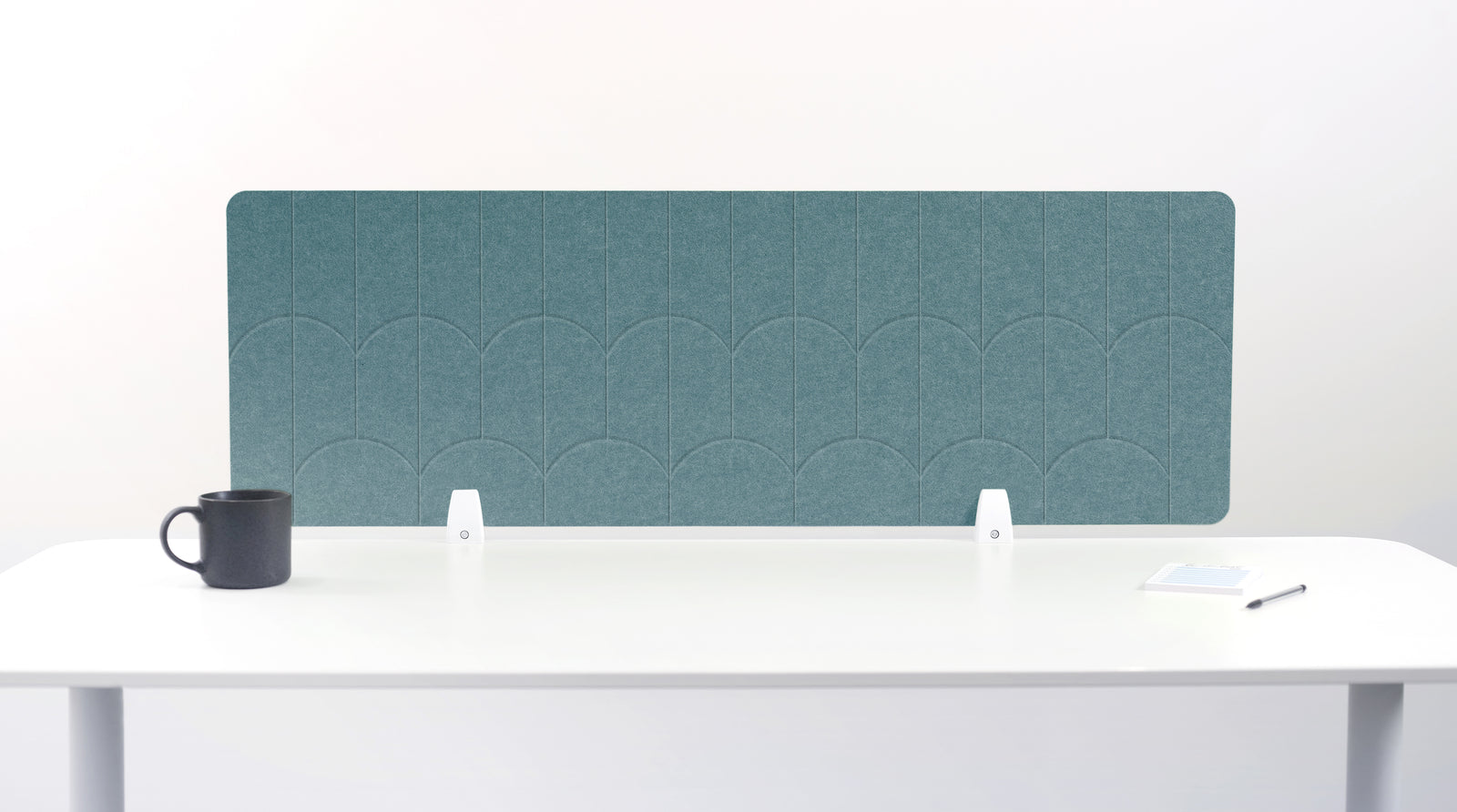 A teal blue felt desk divider with a scalloped wave pattern, mounted on a white desk with a black mug on the left and a small notepad on the right.