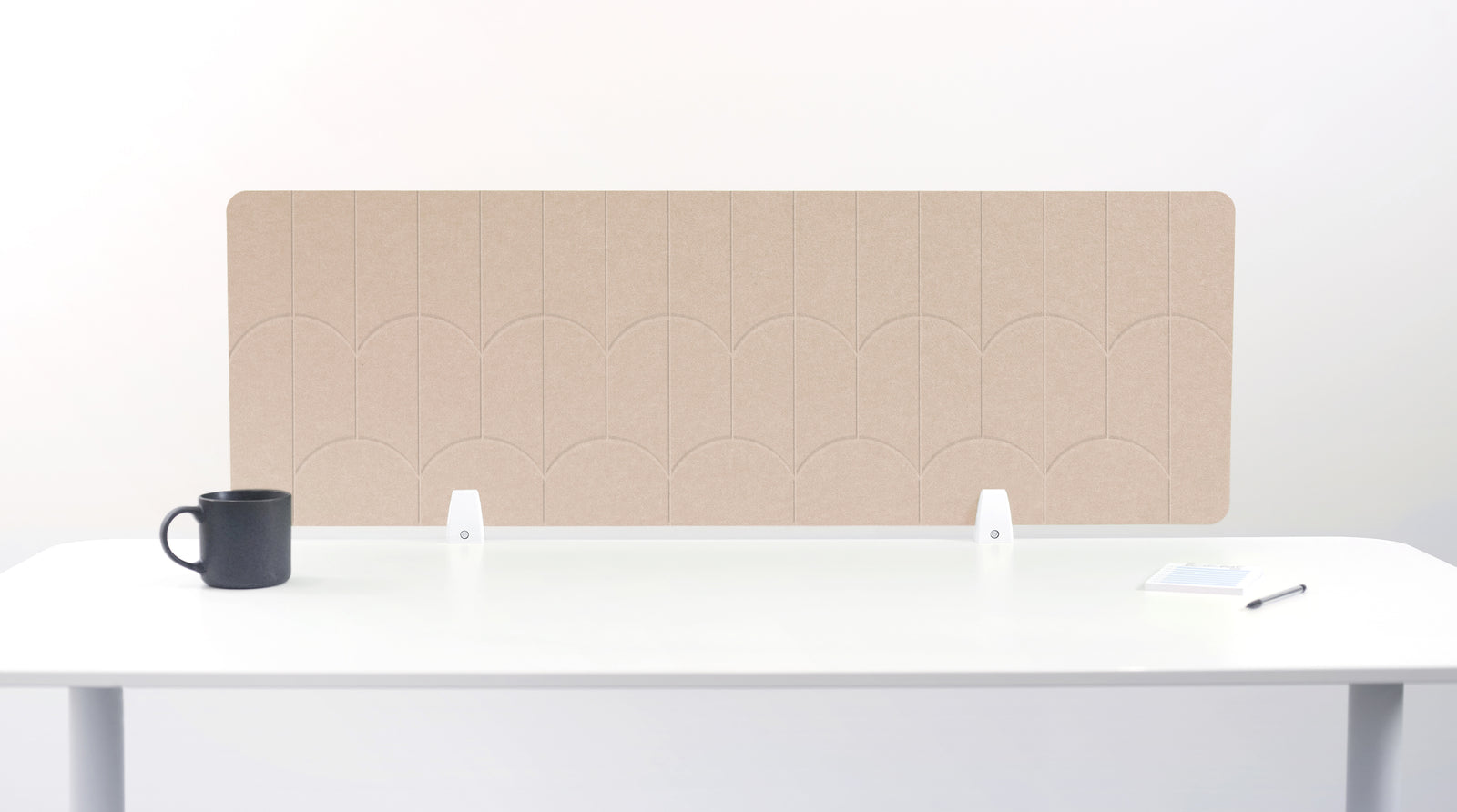 A tan felt desk divider with a scalloped wave pattern, mounted on a white desk with a black mug on the left and a small notepad on the right.