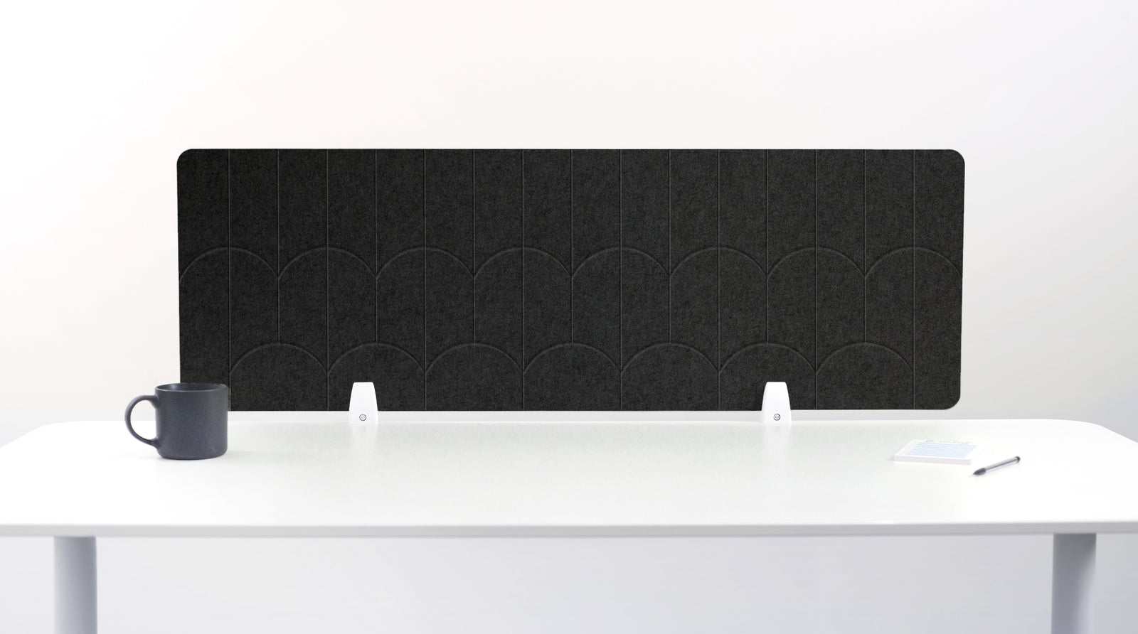 A dark gray felt desk divider with a scalloped wave pattern, mounted on a white desk with a black mug on the left and a small notepad on the right.