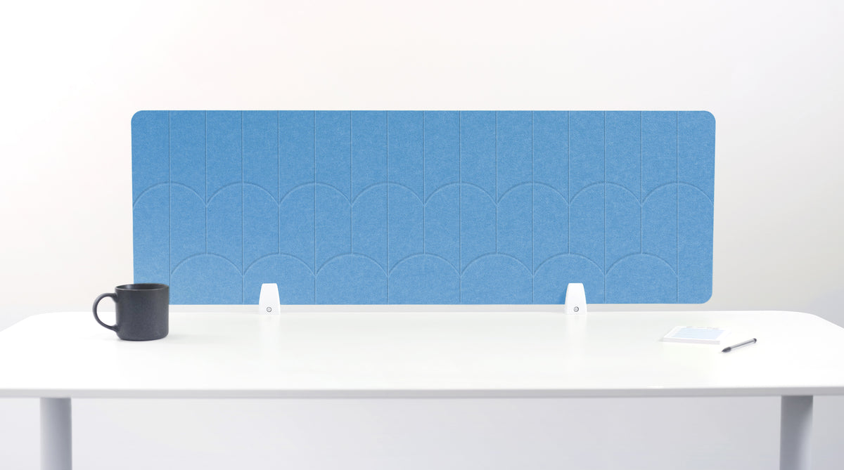 A light blue felt desk divider with a scalloped wave pattern, mounted on a white desk with a black mug on the left and a small notepad on the right.