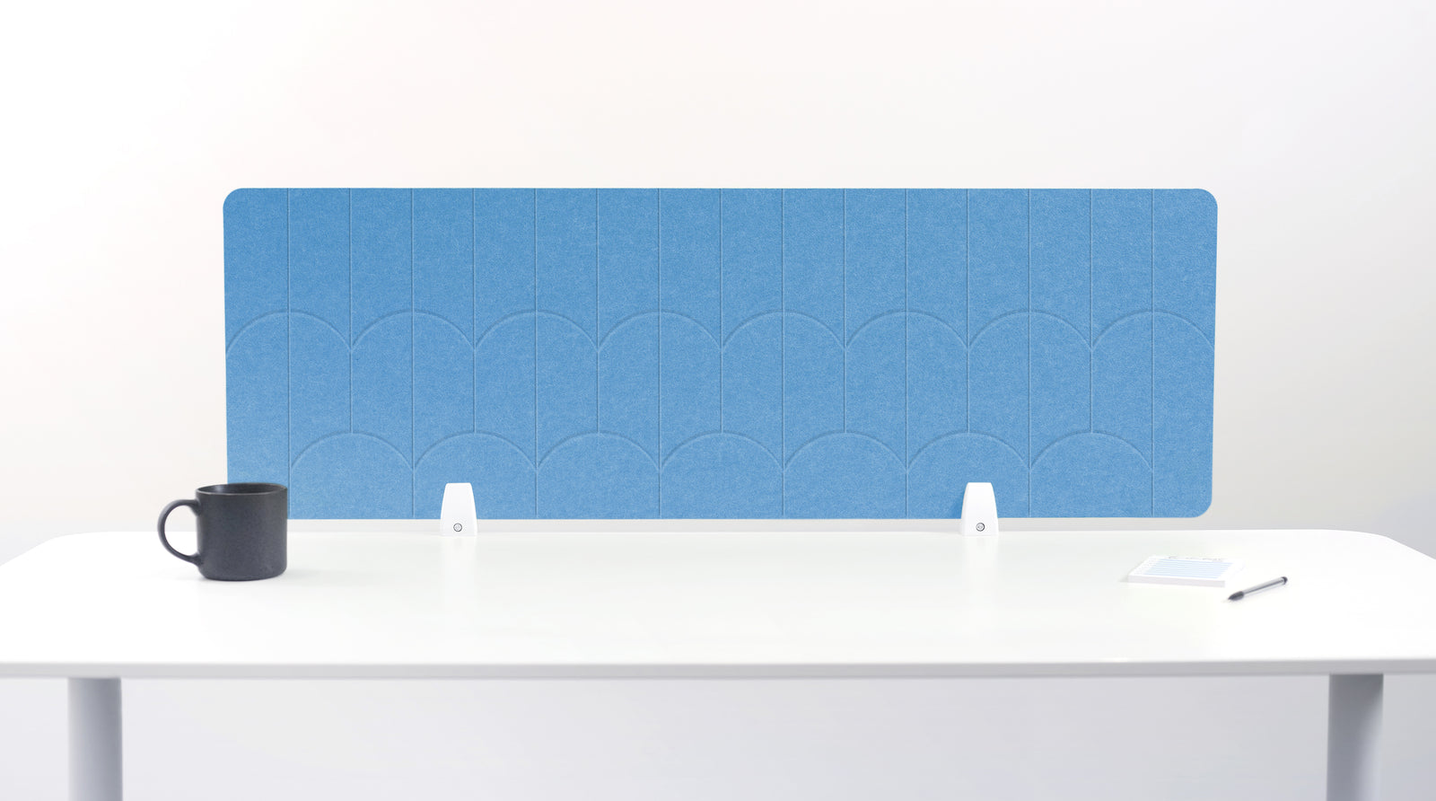 A light blue felt desk divider with a scalloped wave pattern, mounted on a white desk with a black mug on the left and a small notepad on the right.