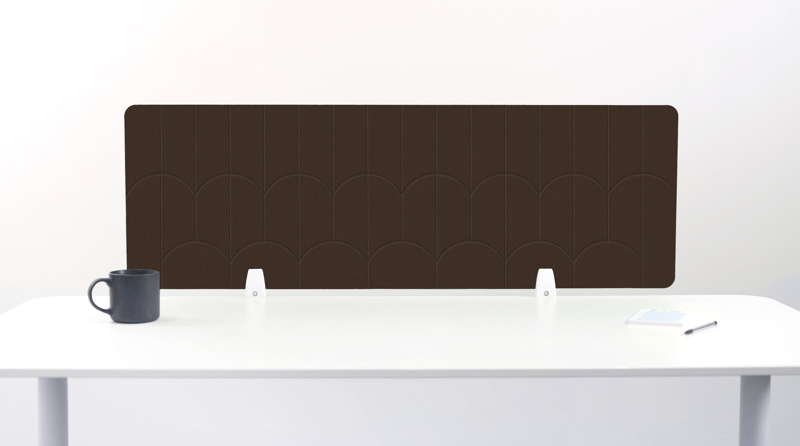A dark brown felt desk divider with a scalloped wave pattern, mounted on a white desk with a black mug on the left and a small notepad on the right.