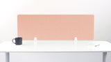A salmon/light pink felt desk divider with a scalloped wave pattern, mounted on a white desk with a black mug on the left and a small notepad on the right.