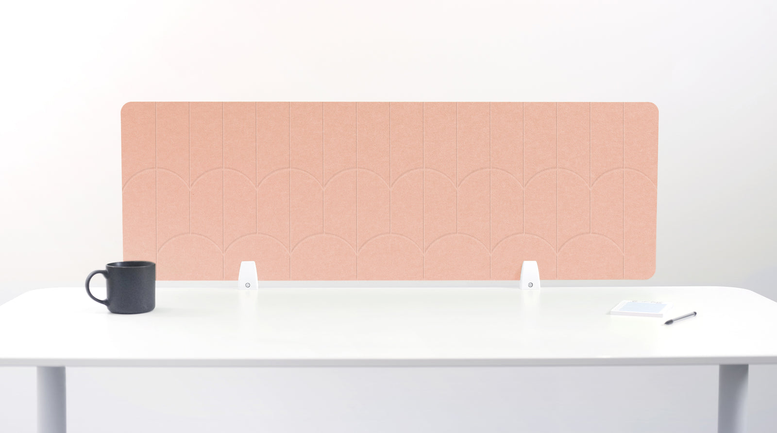 A salmon/light pink felt desk divider with a scalloped wave pattern, mounted on a white desk with a black mug on the left and a small notepad on the right.