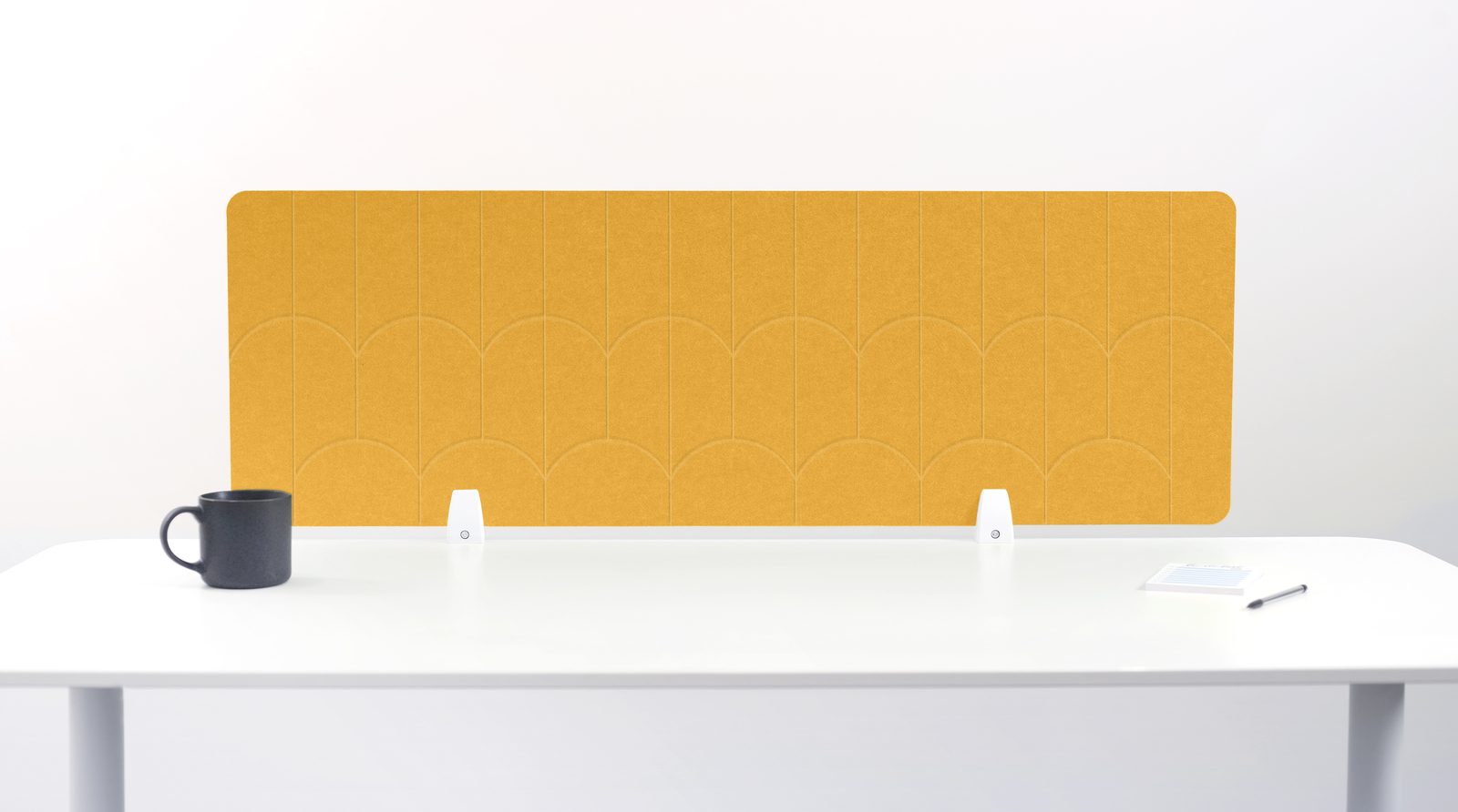 A yellow felt desk divider with a scalloped wave pattern, mounted on a white desk with a black mug on the left and a small notepad on the right.