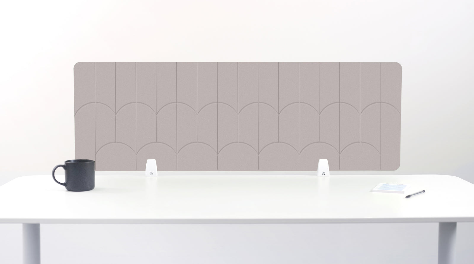 A tan felt desk divider with a scalloped wave pattern, mounted on a white desk with a black mug on the left and a small notepad on the right.