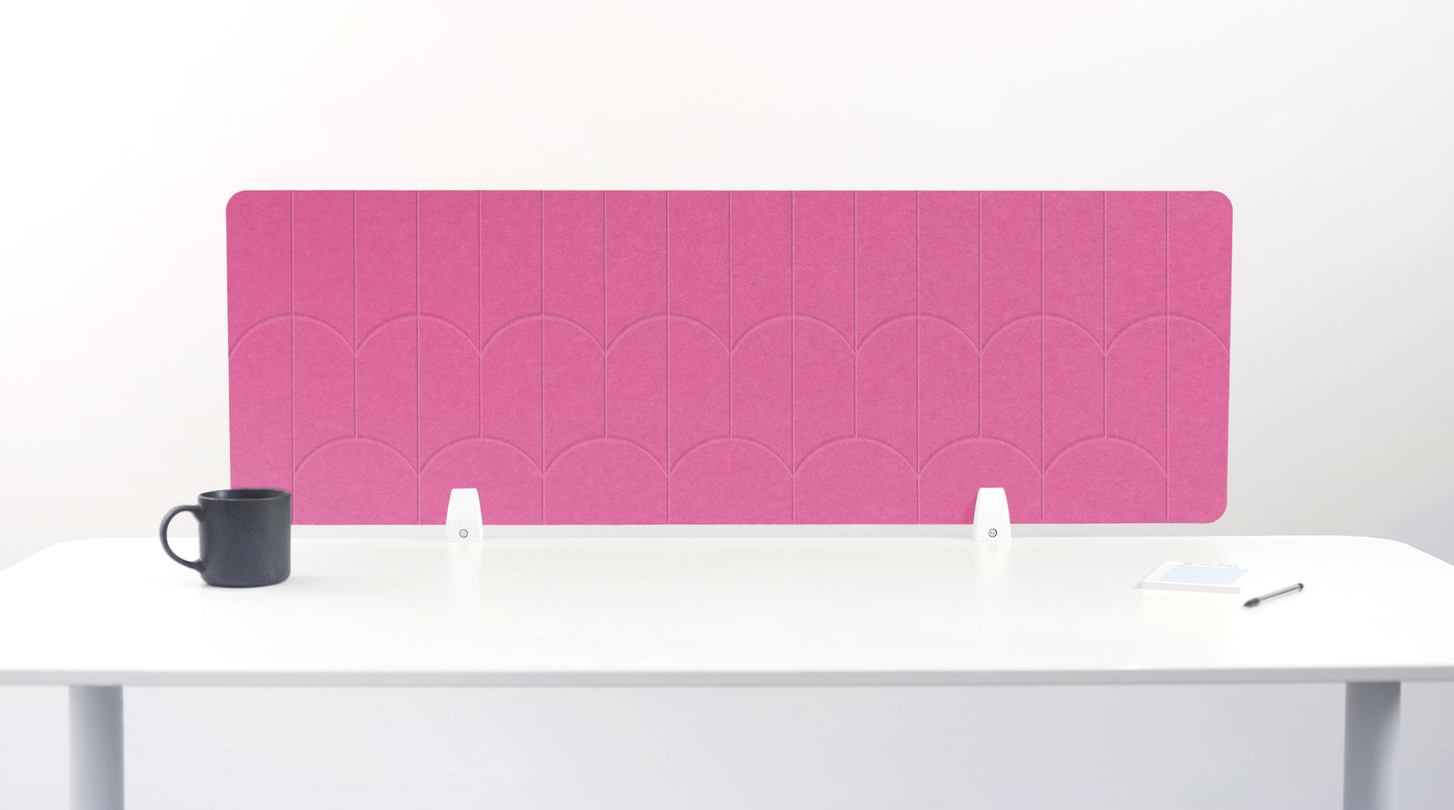 A bright pink felt desk divider with a scalloped wave pattern, mounted on a white desk with a black mug on the left and a small notepad on the right.