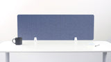A gray/blue felt desk divider with a scalloped wave pattern, mounted on a white desk with a black mug on the left and a small notepad on the right.