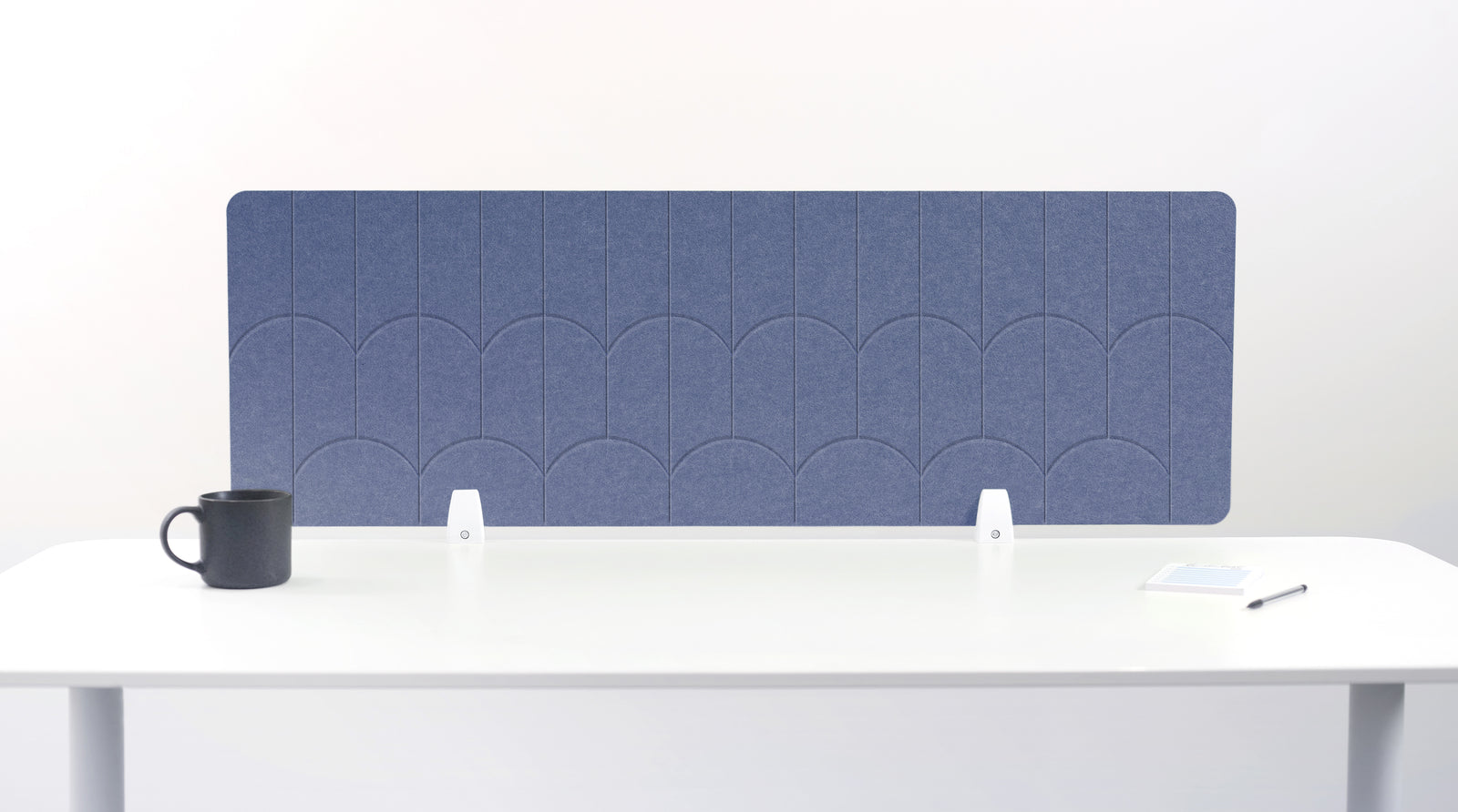 A gray/blue felt desk divider with a scalloped wave pattern, mounted on a white desk with a black mug on the left and a small notepad on the right.