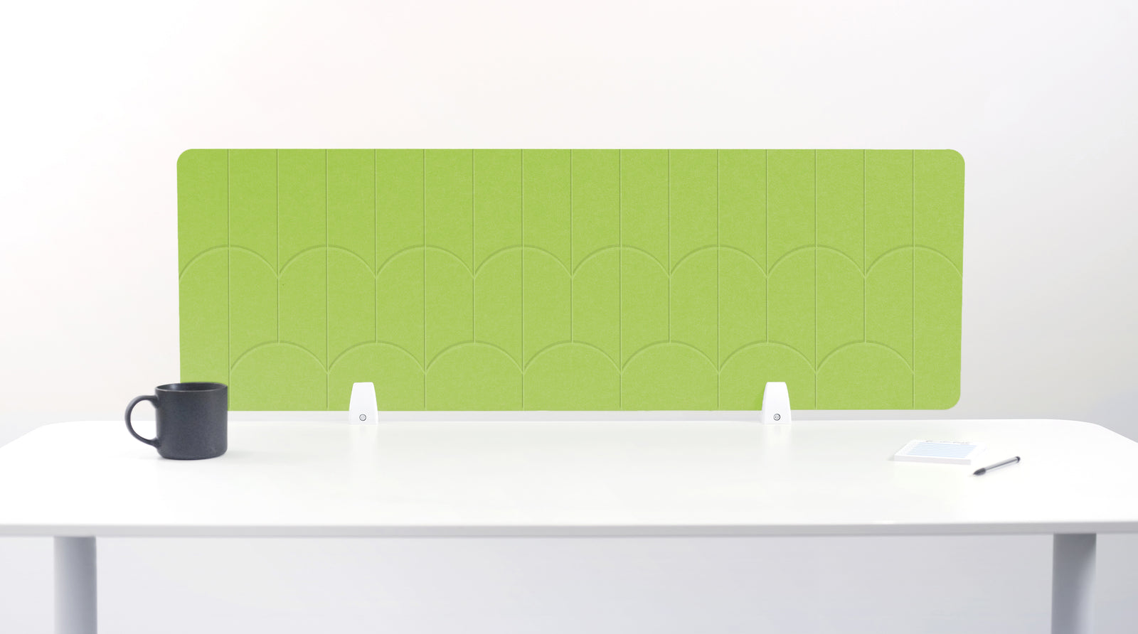 A bright green acoustic desk divider with a scalloped pattern, mounted on a white desk with a black mug on the left and a small notepad on the right.