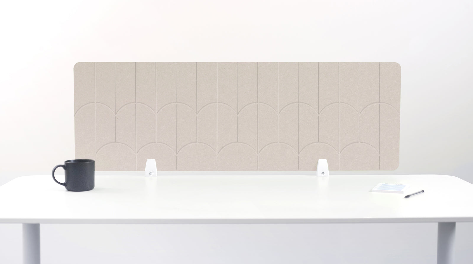 A beige felt desk divider with a scalloped wave pattern, mounted on a white desk with a black mug on the left and a small notepad on the right.