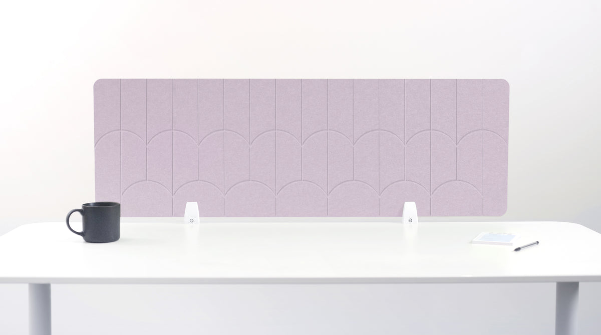 A lilac pink colored felt desk divider with a scalloped wave pattern, mounted on a white desk with a black mug on the left and a small notepad on the right.