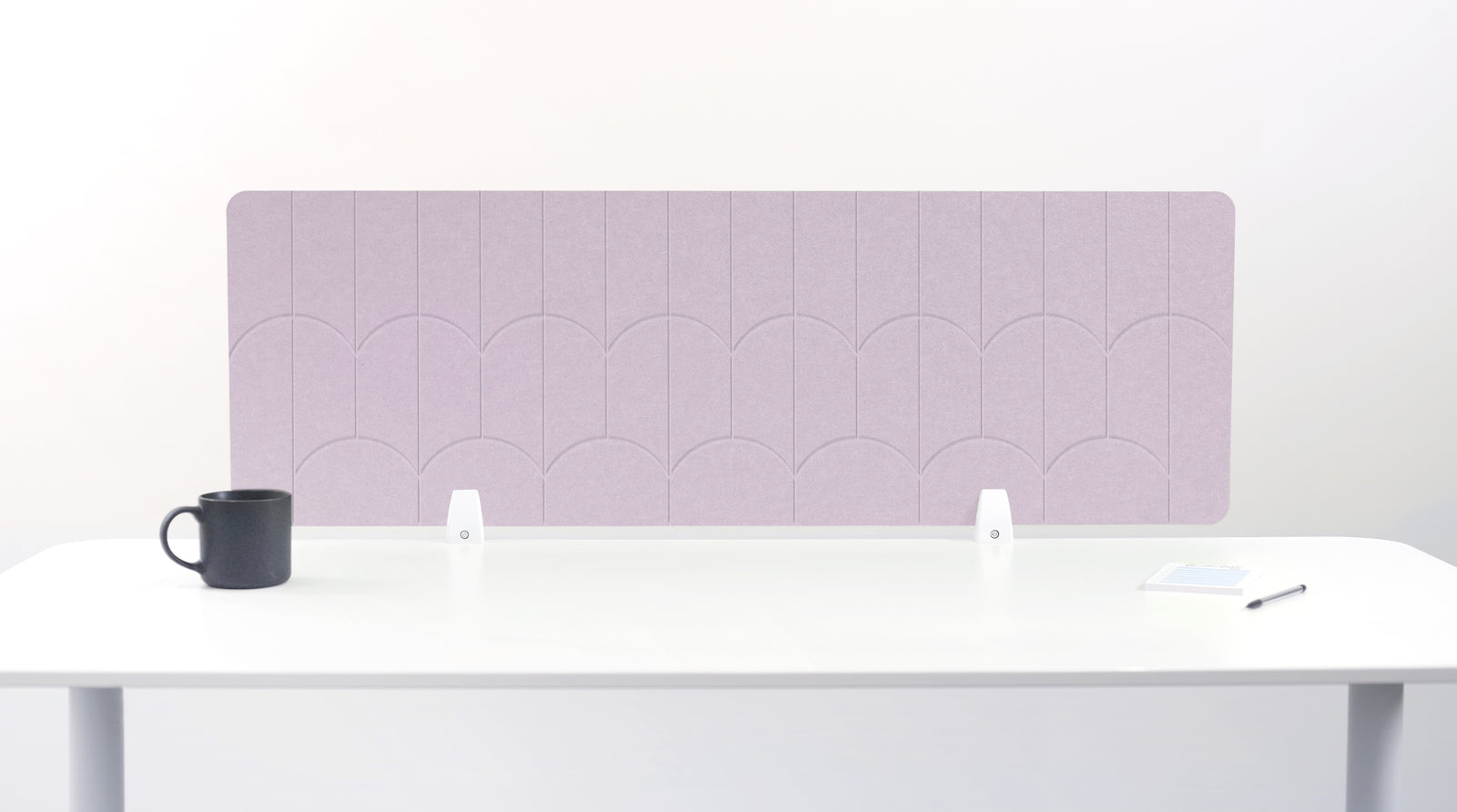 A lilac pink colored felt desk divider with a scalloped wave pattern, mounted on a white desk with a black mug on the left and a small notepad on the right.