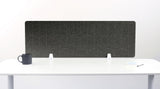 A gray felt desk divider with a scalloped wave pattern, mounted on a white desk with a black mug on the left and a small notepad on the right.
