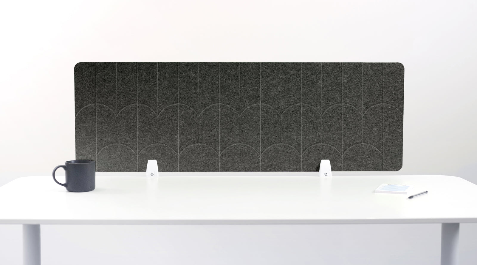A gray felt desk divider with a scalloped wave pattern, mounted on a white desk with a black mug on the left and a small notepad on the right.