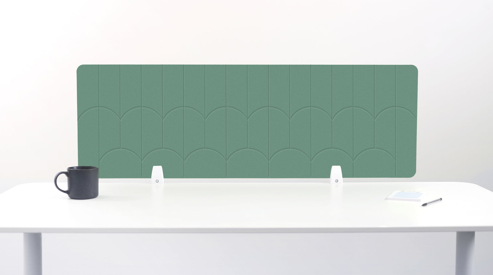 A mint green felt desk divider with a scalloped wave pattern, mounted on a white desk with a black mug on the left and a small notepad on the right.