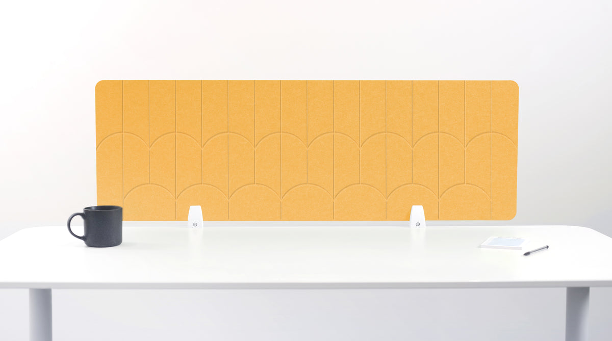 A mustard yellow felt desk divider with a scalloped wave pattern, mounted on a white desk with a black mug on the left and a small notepad on the right.