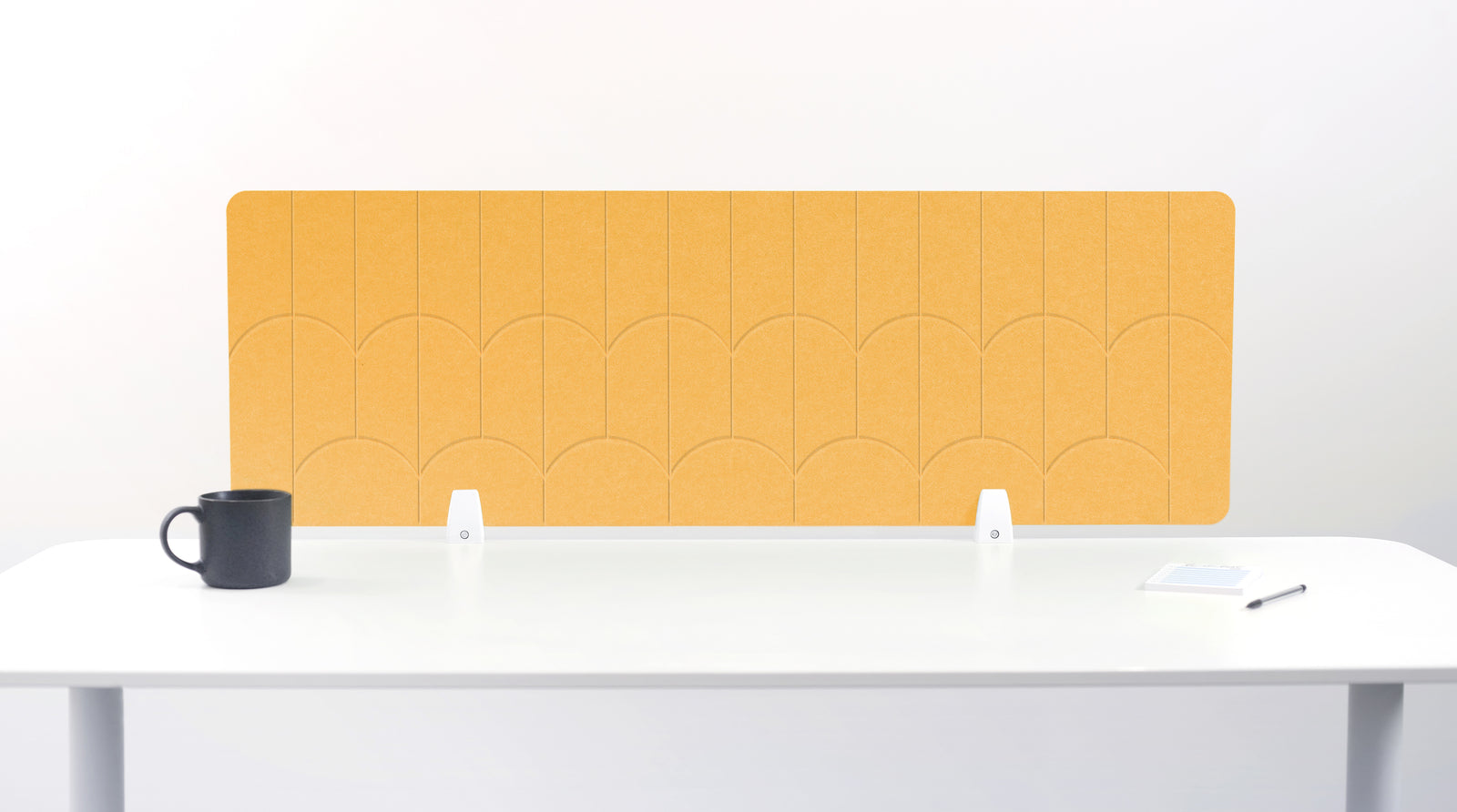 A mustard yellow felt desk divider with a scalloped wave pattern, mounted on a white desk with a black mug on the left and a small notepad on the right.