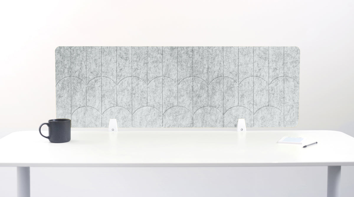 A nickel colored felt desk divider with a scalloped wave pattern, mounted on a white desk with a black mug on the left and a small notepad on the right.