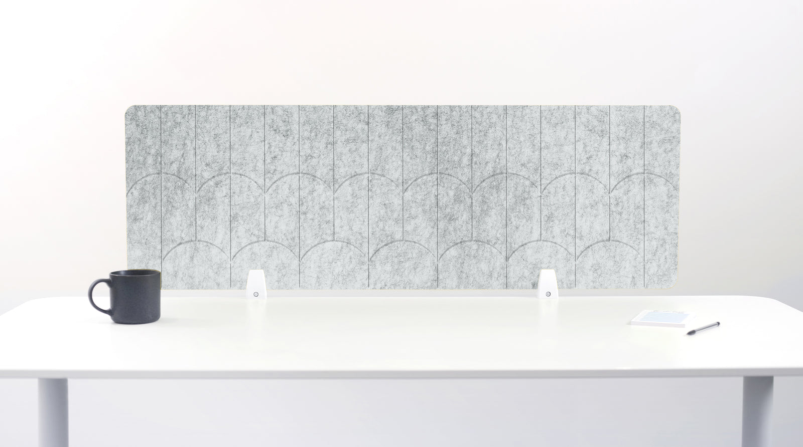 A nickel colored felt desk divider with a scalloped wave pattern, mounted on a white desk with a black mug on the left and a small notepad on the right.