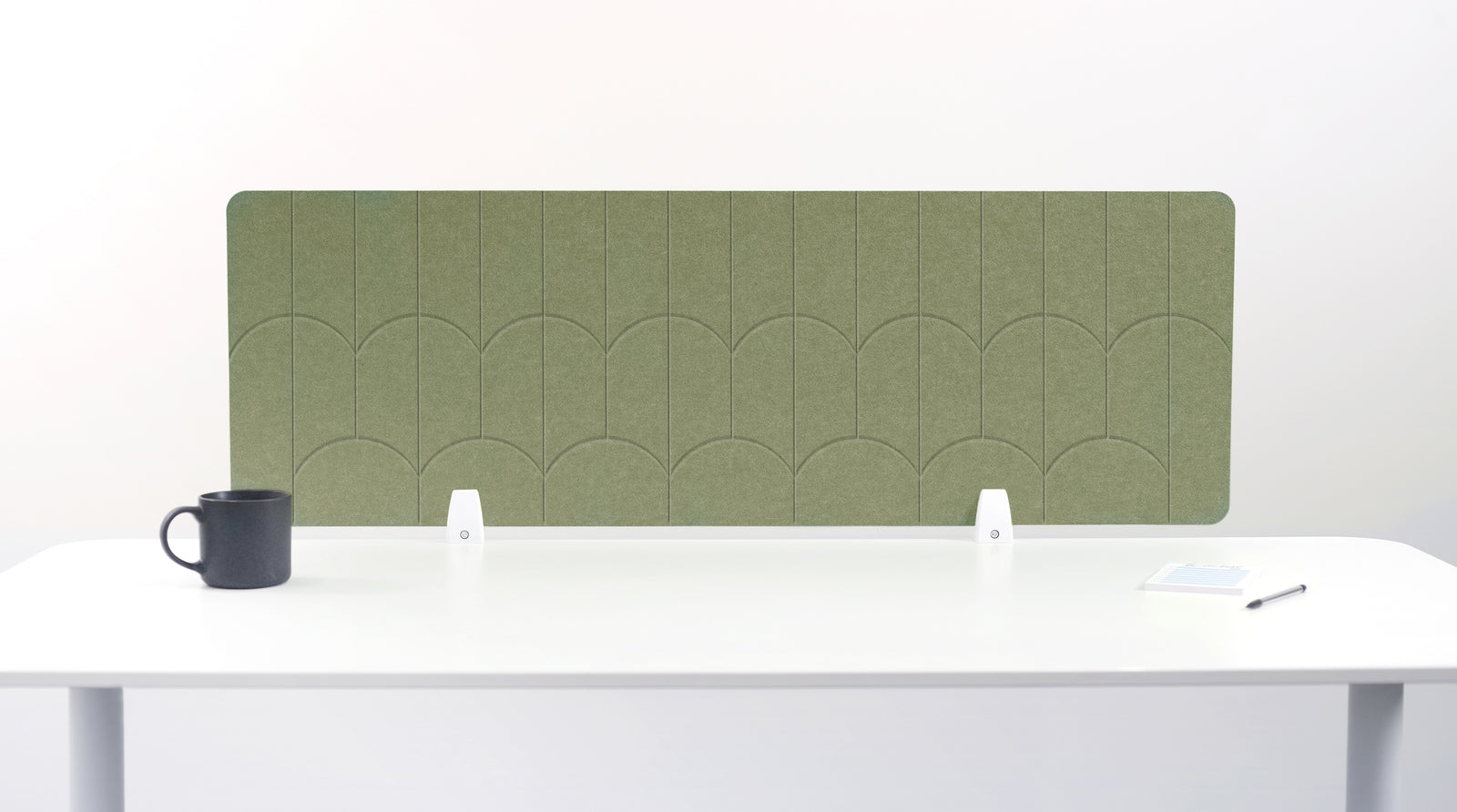 An olive green felt desk divider with a scalloped wave pattern, mounted on a white desk with a black mug on the left and a small notepad on the right.