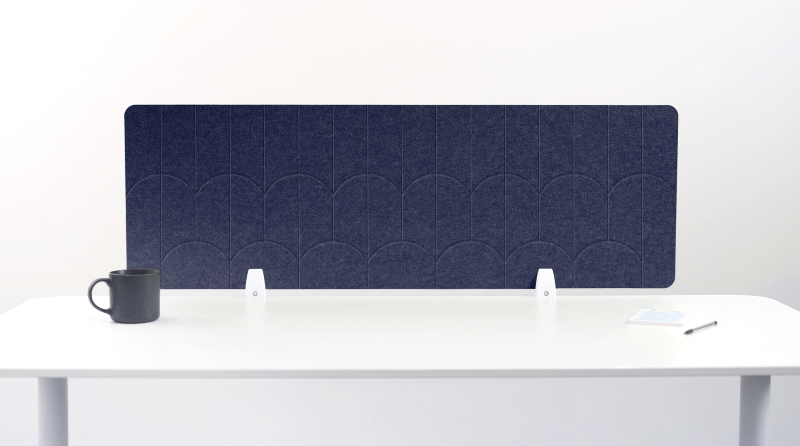 A navy blue felt desk divider with a scalloped wave pattern, mounted on a white desk with a black mug on the left and a small notepad on the right.