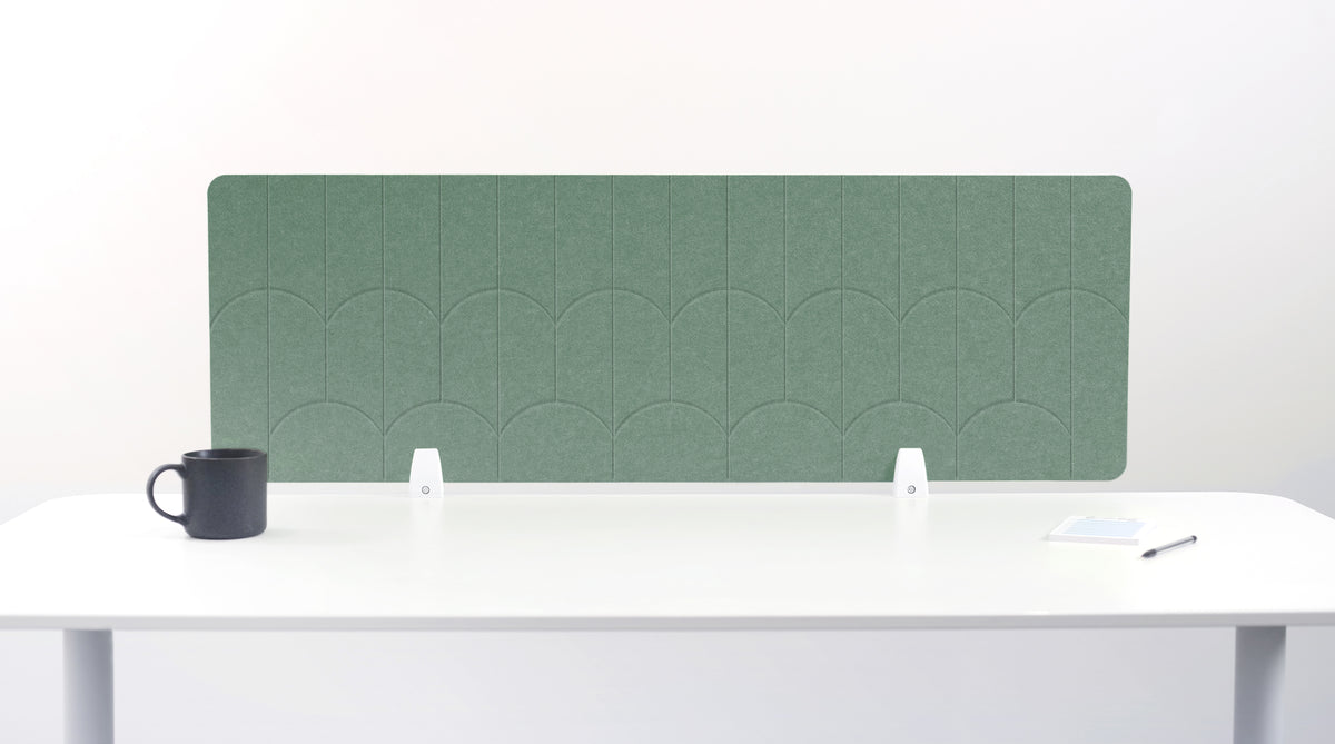 A green felt desk divider with a scalloped wave pattern, mounted on a white desk with a black mug on the left and a small notepad on the right.