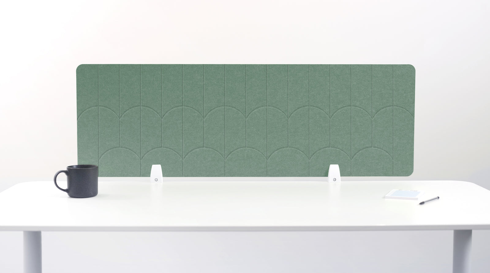 A green felt desk divider with a scalloped wave pattern, mounted on a white desk with a black mug on the left and a small notepad on the right.