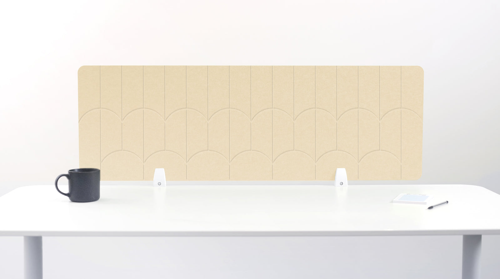 A light tan felt desk divider with a scalloped wave pattern, mounted on a white desk with a black mug on the left and a small notepad on the right.