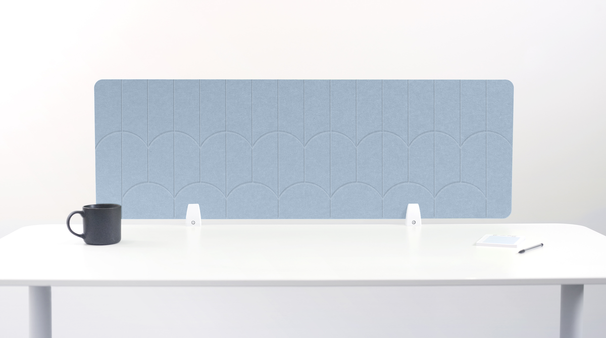 A light blue felt desk divider with a scalloped arch pattern, mounted on a white desk with a black mug, small white objects, and a notepad visible.