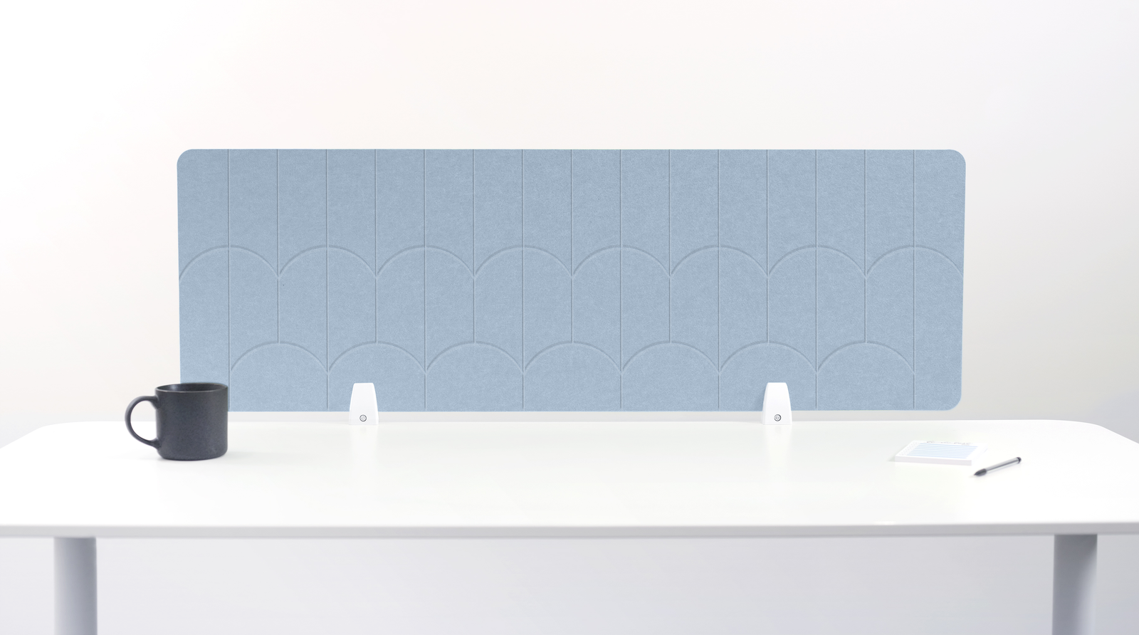 A light blue felt desk divider with a scalloped arch pattern, mounted on a white desk with a black mug, small white objects, and a notepad visible.