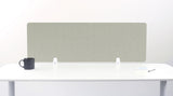 A sage green felt desk divider with a scalloped wave pattern, mounted on a white desk with a black mug on the left and a small notepad on the right.