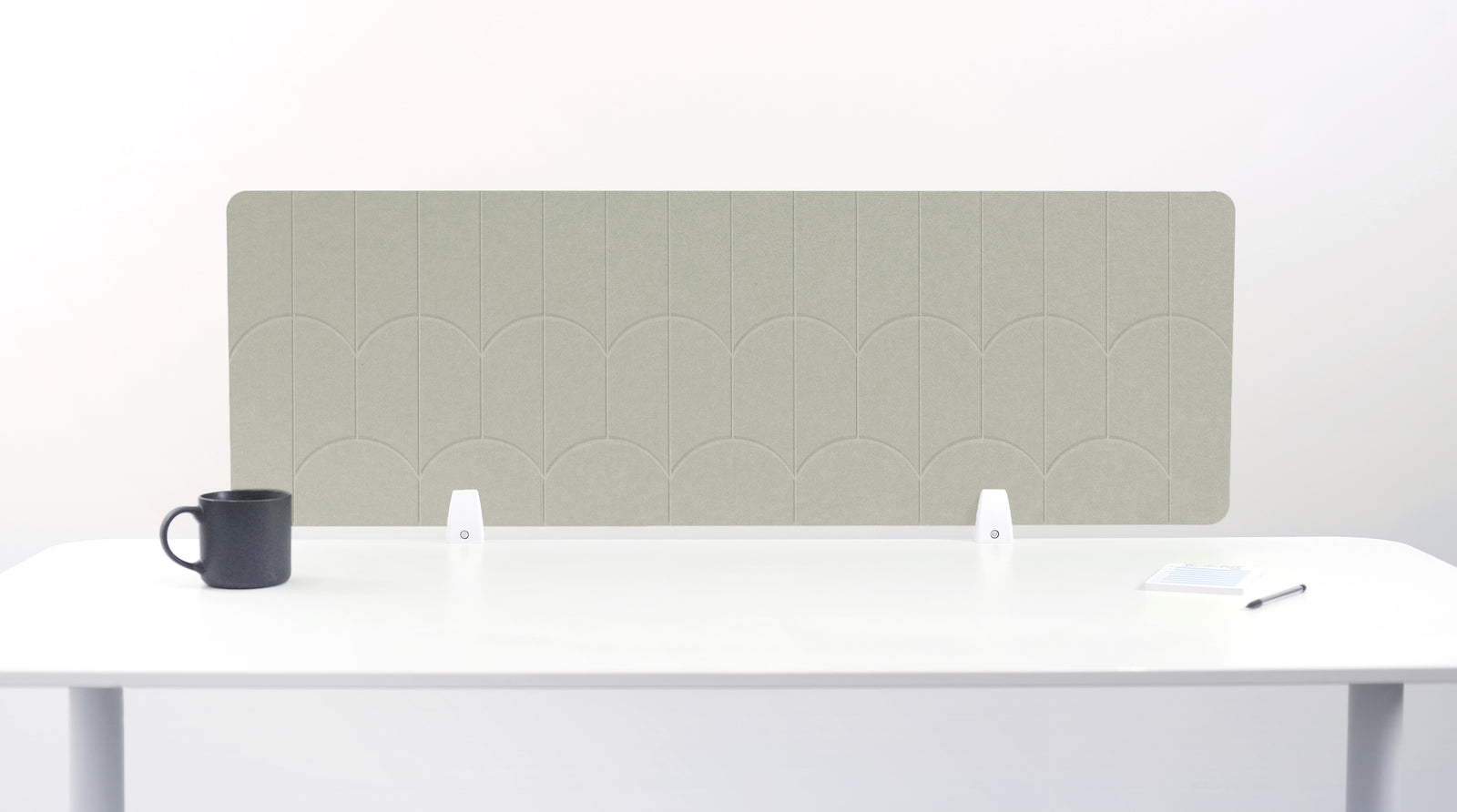 A sage green felt desk divider with a scalloped wave pattern, mounted on a white desk with a black mug on the left and a small notepad on the right.