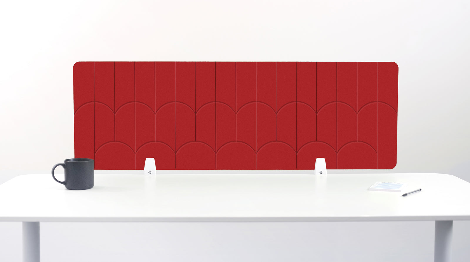 A red desk divider panel with a scalloped pattern, mounted on a white desk with a black mug on the left and a small notepad on the right.