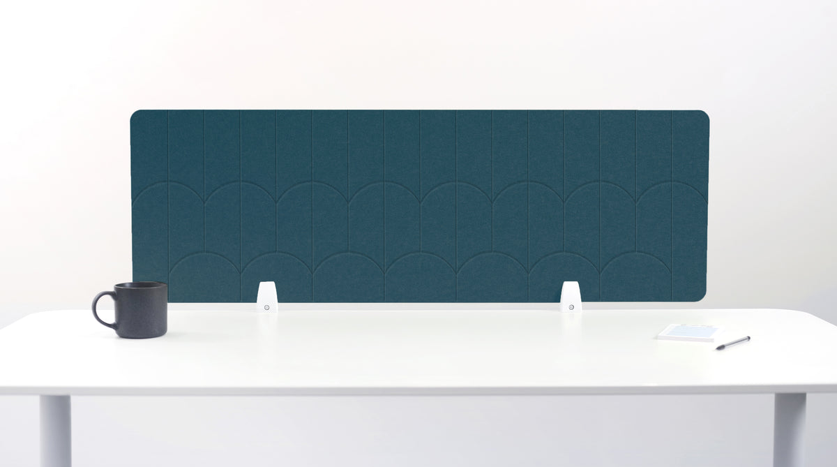A slate blue felt desk divider with a scalloped wave pattern, mounted on a white desk with a black mug on the left and a small notepad on the right.