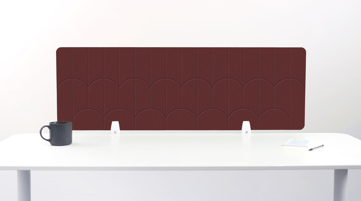 A dark reddish-brown desk divider panel with a scalloped pattern, mounted on a white desk with a black mug on the left and a small notepad on the right.