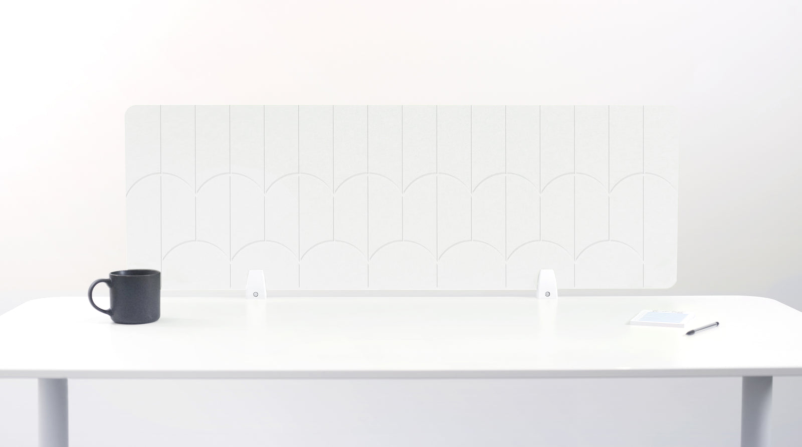 A white felt desk divider with a scalloped wave pattern, mounted on a white desk with a black mug on the left and a small notepad on the right.
