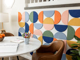 Felt right wall art design by a kitchen table in shades of pink, blue, green, and orange felt half circle tiles with some art supplies on the table.