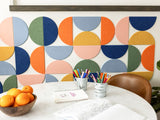 Felt right wall art design by a kitchen table in shades of pink, blue, green, and orange felt half circle tiles with some art supplies on the table.
