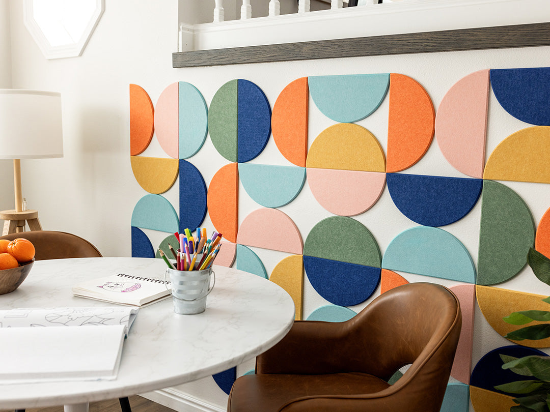 A Felt Right wall art design using half circles in colors orange, pink, blue, yellow, and green creating a geometric pattern on a white wall in front of a table with two chairs and a lamp in the corner.