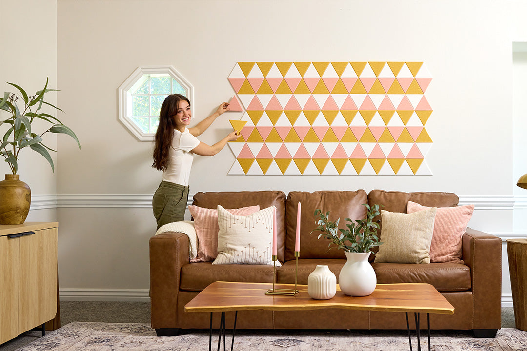 A Felt Right wall art design in shades of yellow pink and white triangles in a living room wall above a couch next to a window.