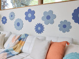 Felt Right wall art design in shades of blue in the shape of flowers on a white wall above a couch with decorative pillows and below a window.