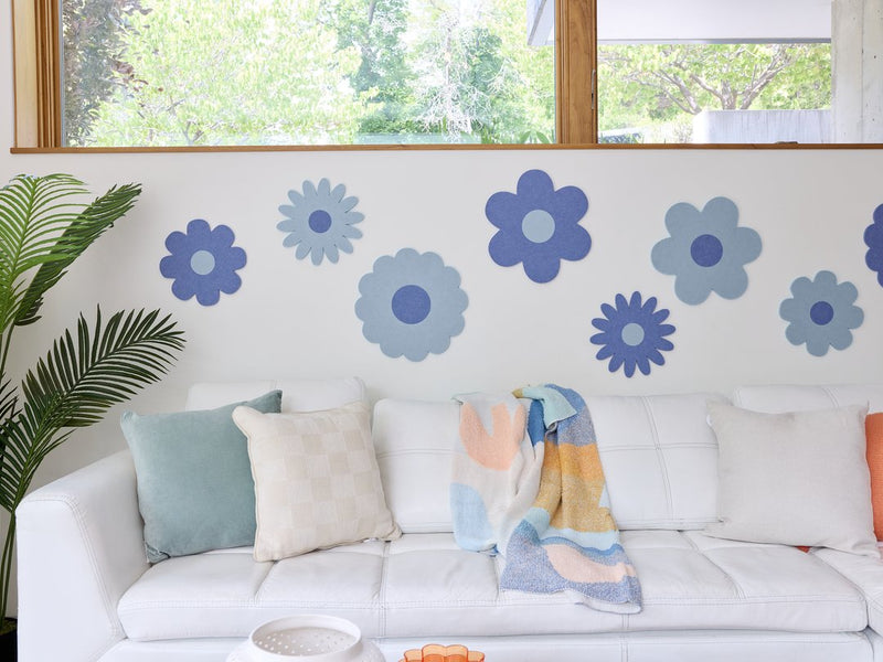 Felt Right wall art design in shades of blue in the shape of flowers on a white wall above a couch and below a window with a house plant in the corner.