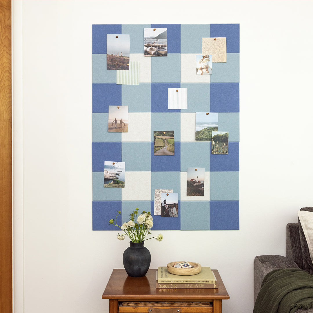 Blue checkered bulletin board with pinned photos on a white wall, next to a wooden side table with a vase and books.