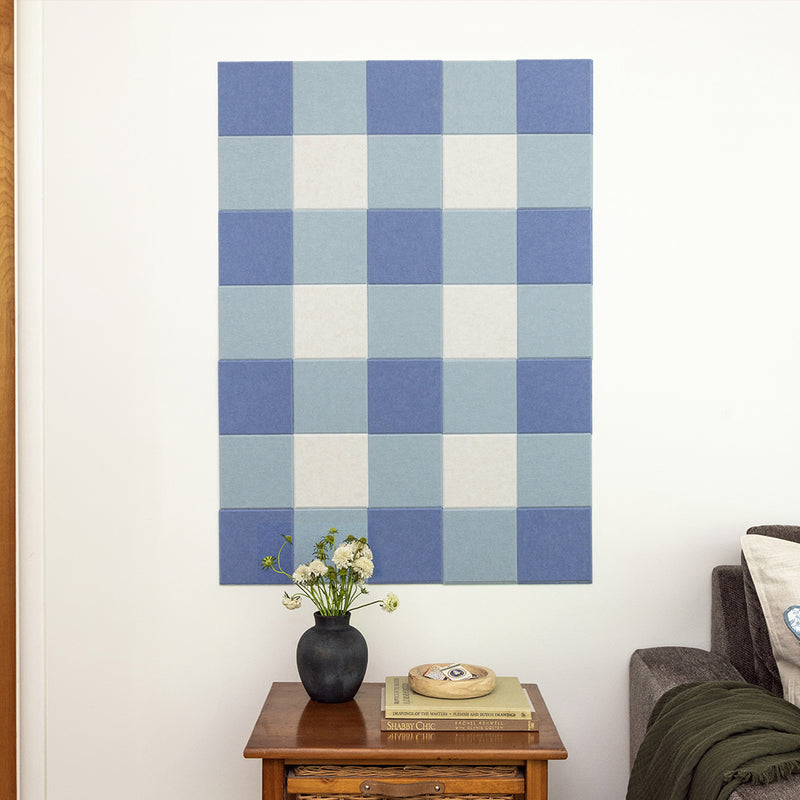 Checkered blue and white wall art on a white wall above a wooden side table with a vase and books.
