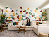 Felt Right wall art design using triangles in shades of blue, red, and yellow creating a geometric pattern on a white wall in a living room with a couch and a woman drinking from a mug and two house plants in the corners.