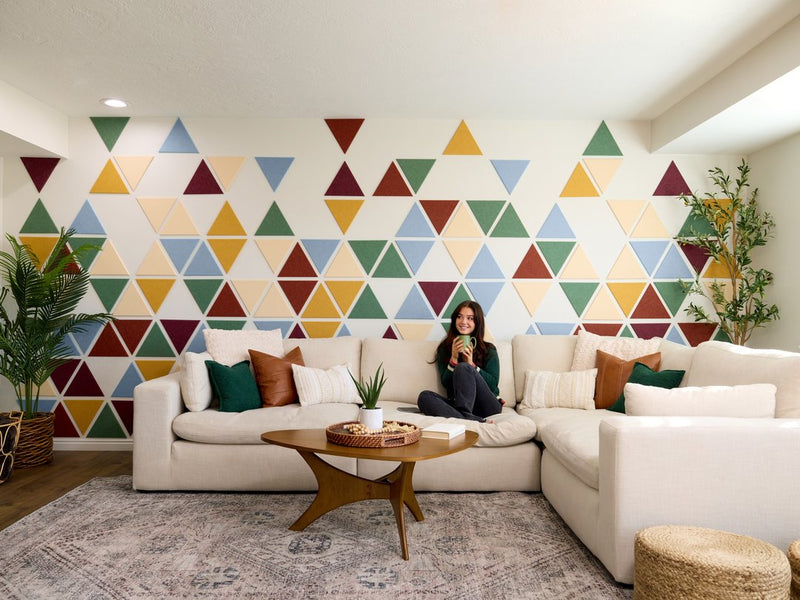 Felt Right wall art design using triangles in shades of blue, red, and yellow creating a geometric pattern on a white wall in a living room with a couch and a woman drinking from a mug and two house plants in the corners.