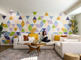 Felt Right wall art design using triangles in shades of yellow, green, blue, and purple on a white wall in a living room space with a woman reading on the couch.