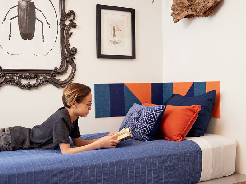 A Felt Right wall art design in shades of blue and orange in a corner of a bedroom above a kids bed and the kid is reading a book on the bed