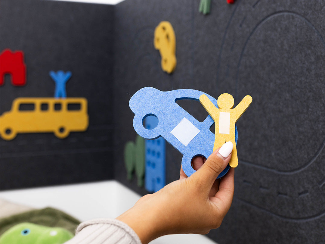 Hand holding a blue car and yellow figure cutout in front of a black board with colorful cutouts.